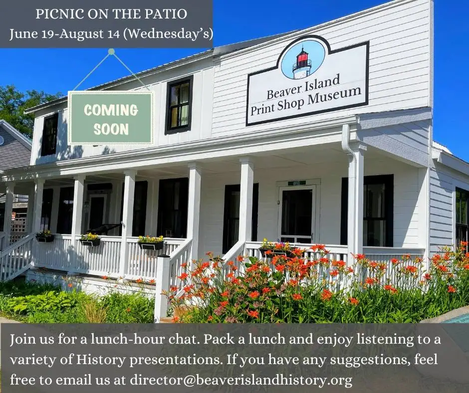 White clapboard building with a covered porch, museum sign, promotional banners, and orange flowers in front.