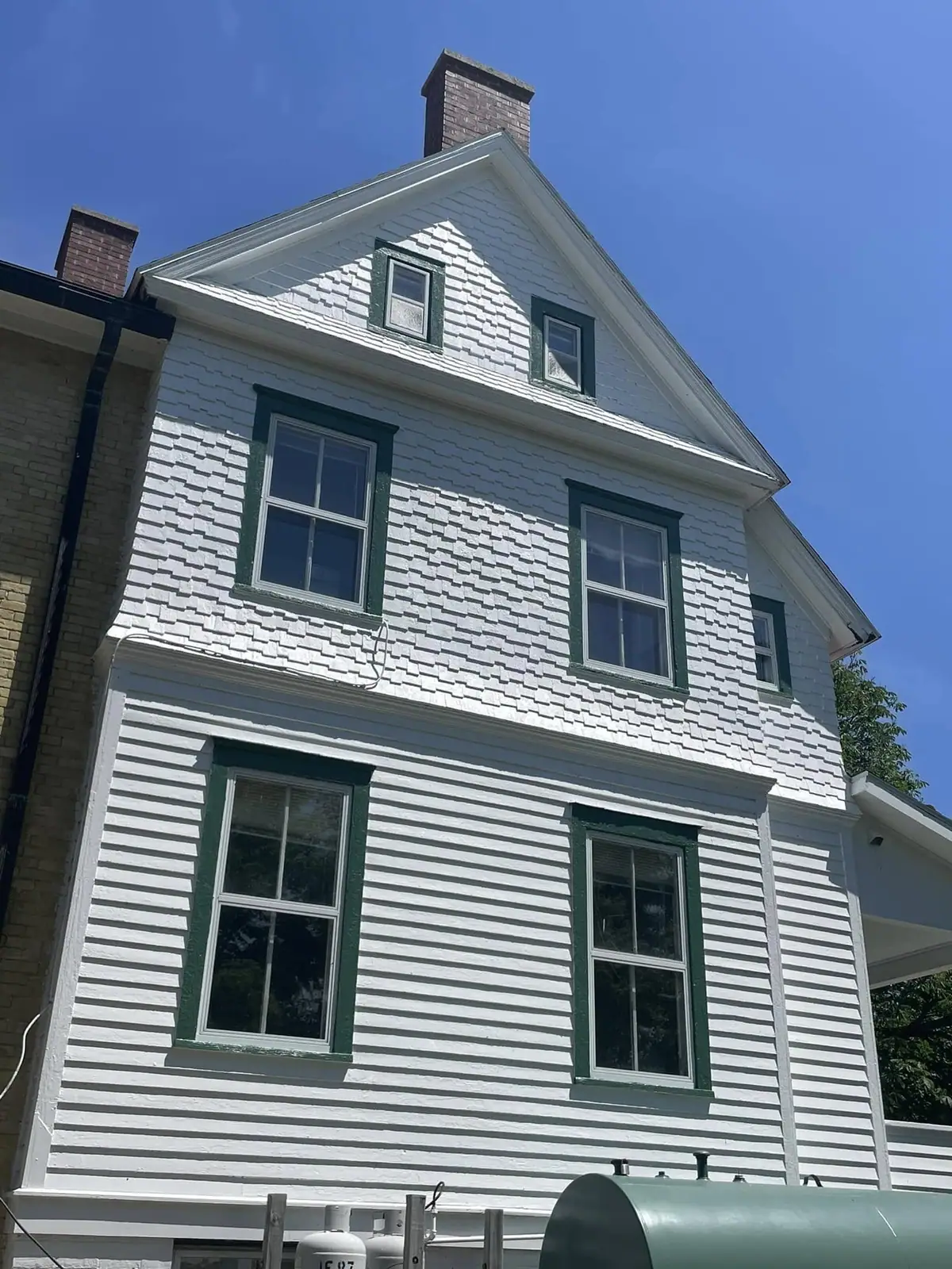 A tall white two-story house with green window trim, shingle siding, dormers, and brick chimneys.