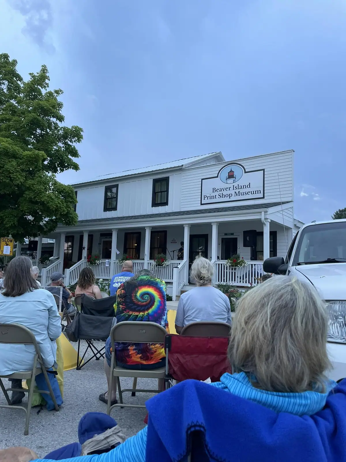 People seated in folding chairs face a white two-story building with a porch and a museum sign.
