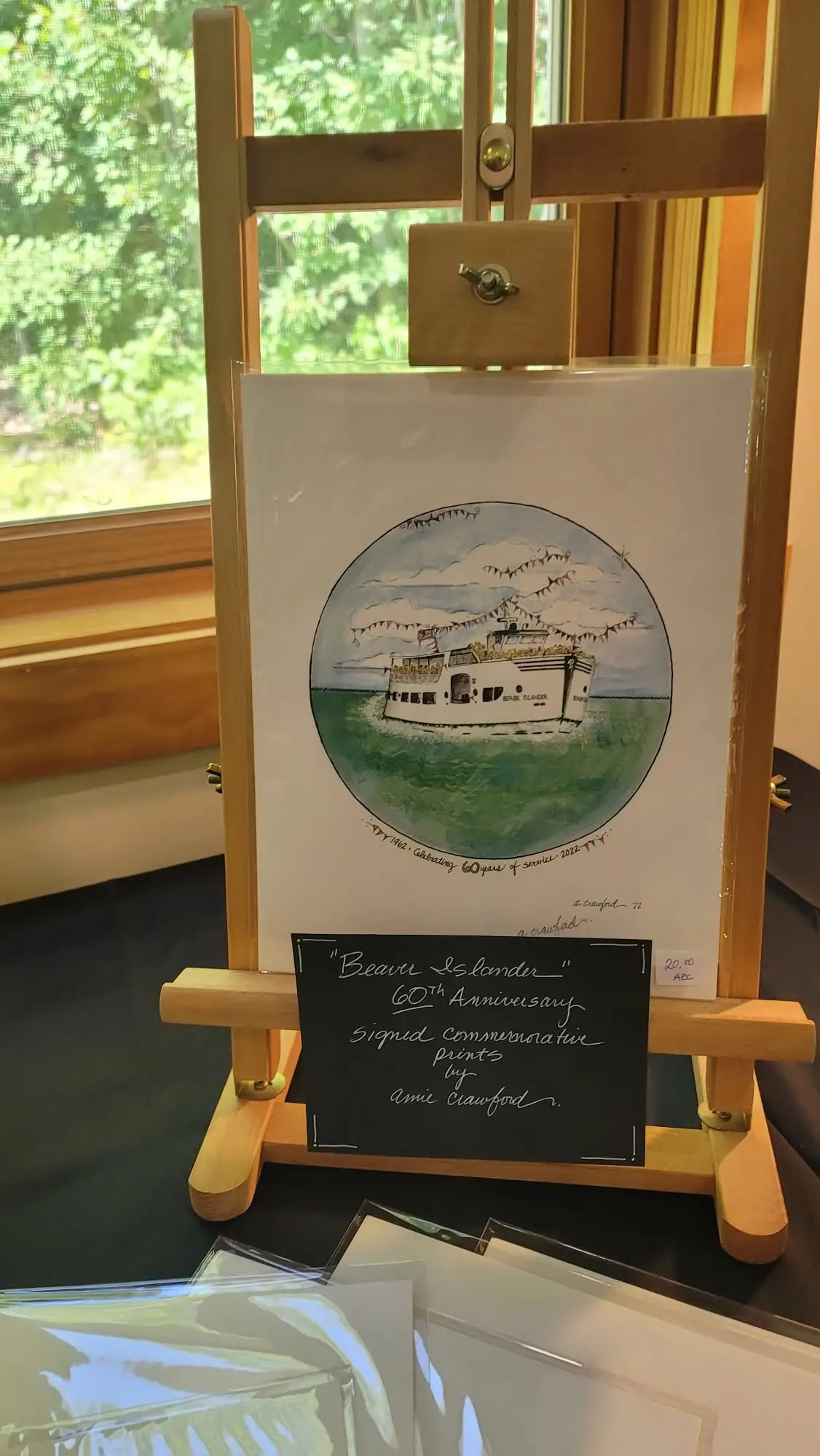 A watercolor ferry print on a wooden easel, with a handwritten anniversary sales sign in front.