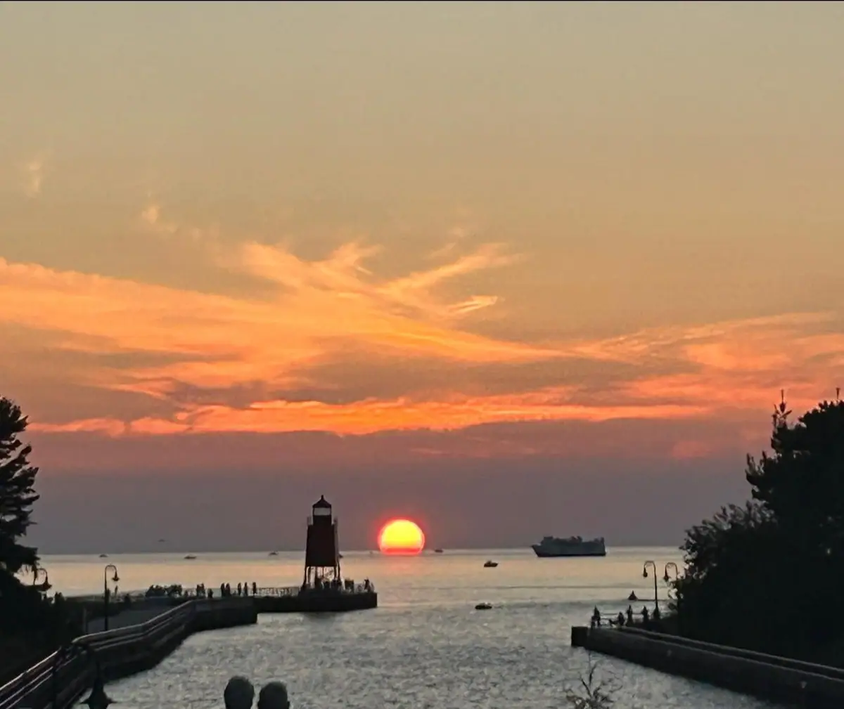 Sunset over a channel with a lighthouse, piers, silhouetted people, and a large vessel offshore.