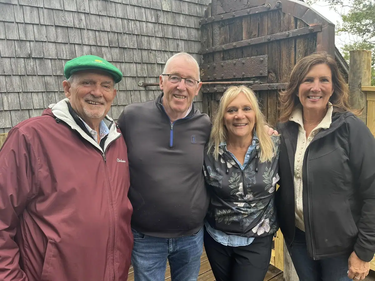 Four smiling adults pose in front of a weathered shingled building and large wooden door.