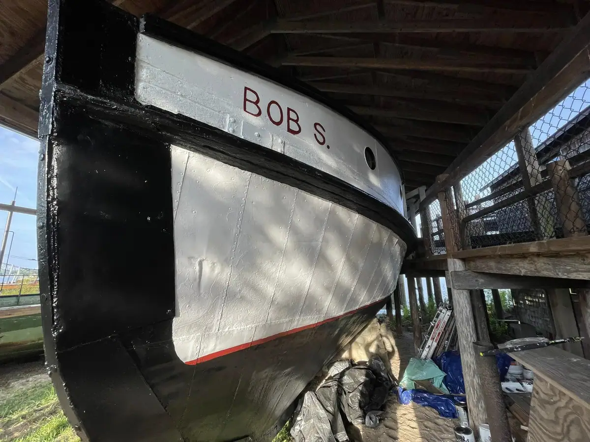 A black-and-white boat hull marked “BOB S.” stands beneath a wooden shelter surrounded by work materials.
