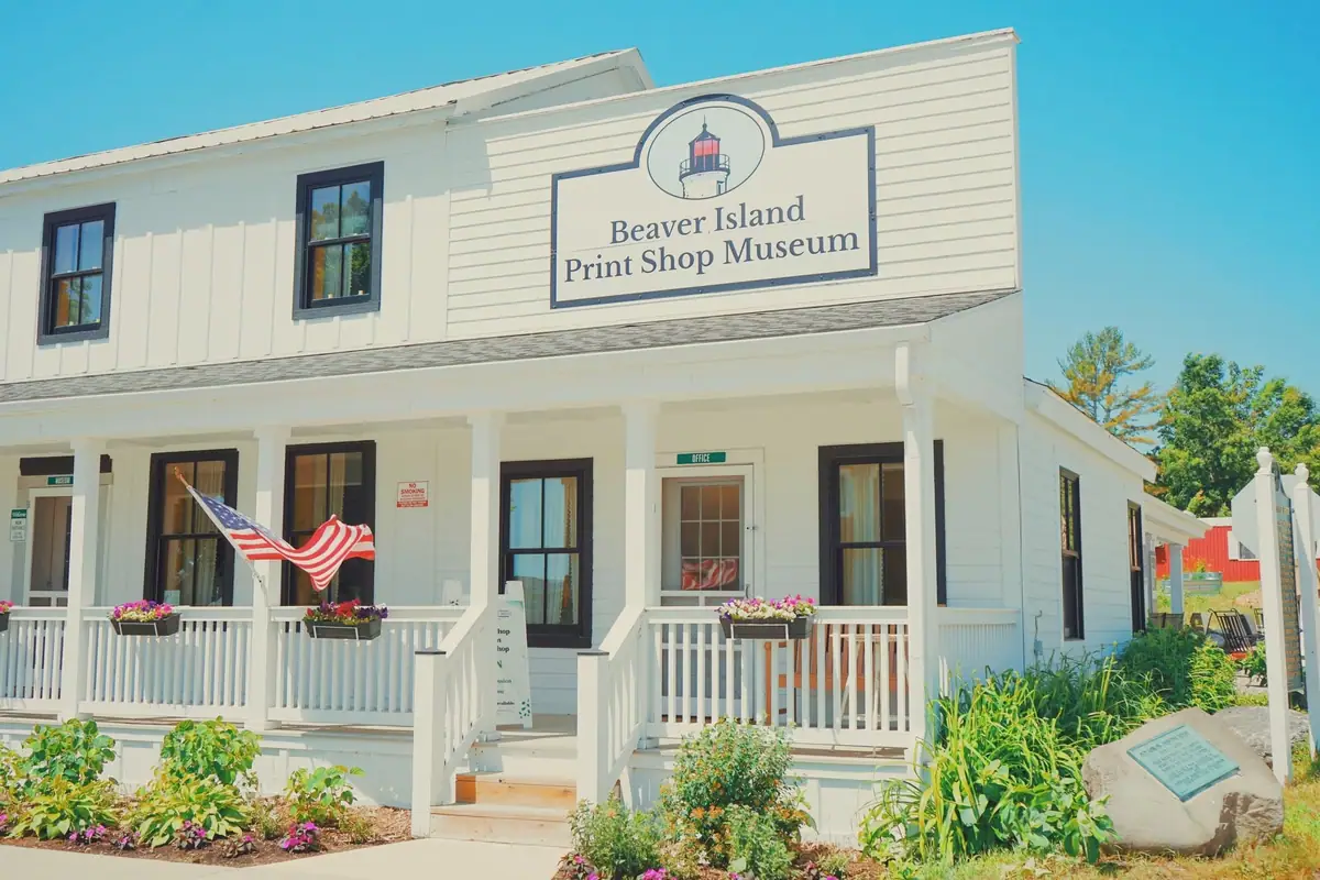 A white wood-sided museum building with a porch, flower boxes, an American flag, and a large sign.