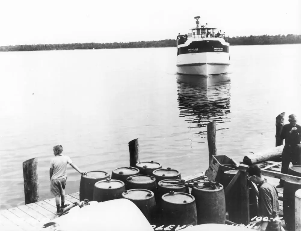 A ferry approaches a wooden dock crowded with barrels, pilings, and several people.