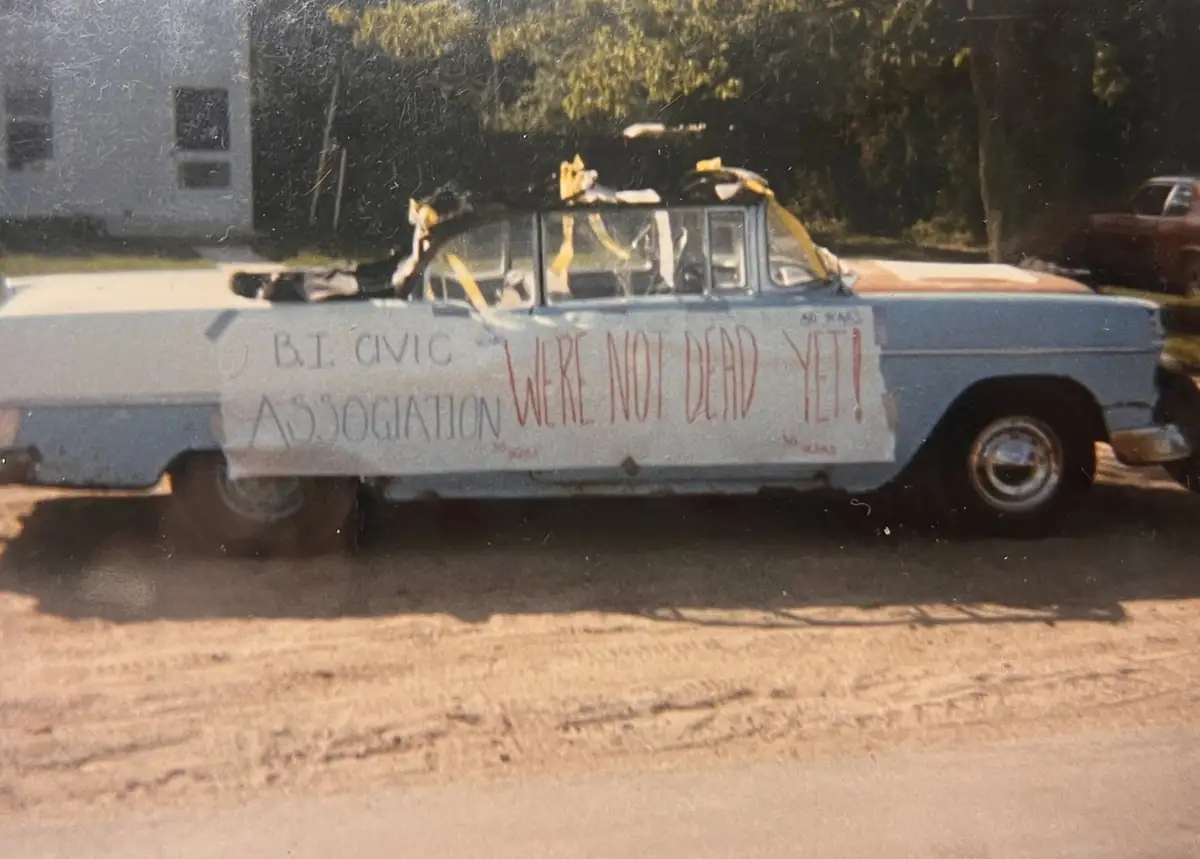 A decorated light blue and white sedan carries a hand-lettered civic association sign beside an unpaved road.