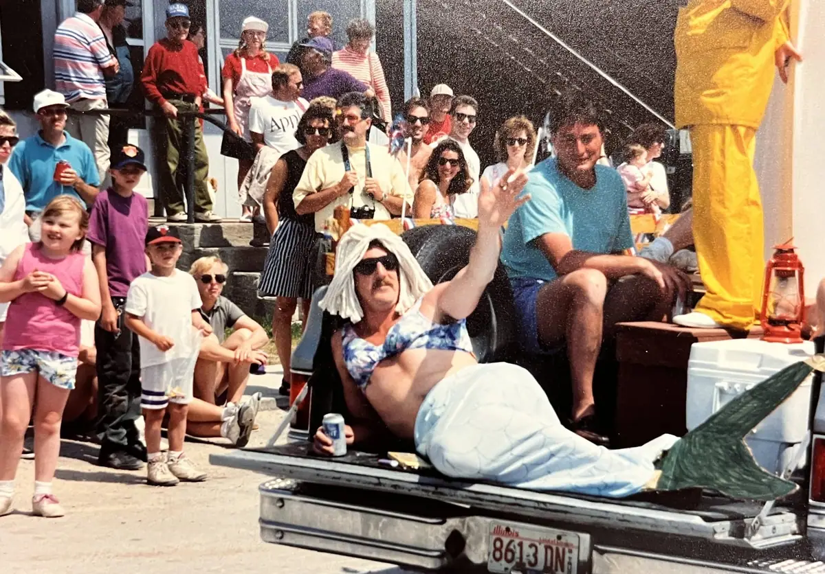 A costumed man in a mermaid tail reclines in a pickup truck while adults and children watch.