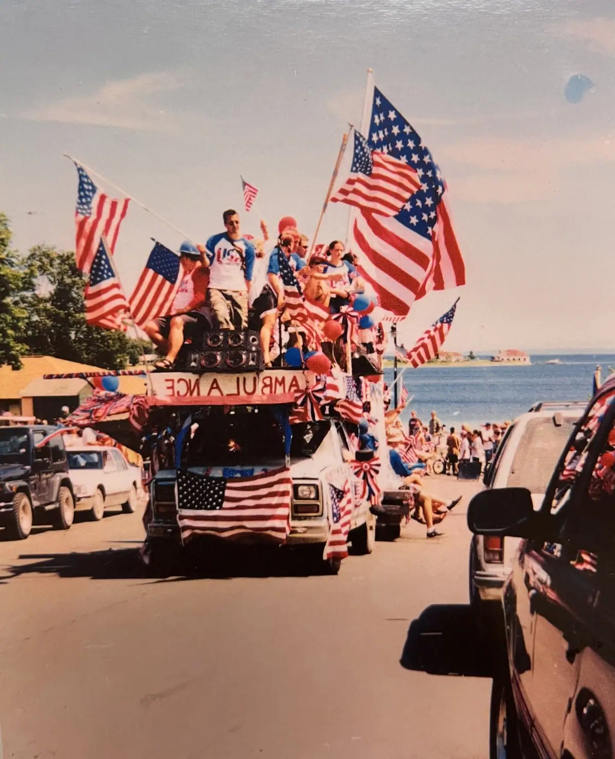 A flag-covered parade vehicle carries people past spectators on a waterfront road.