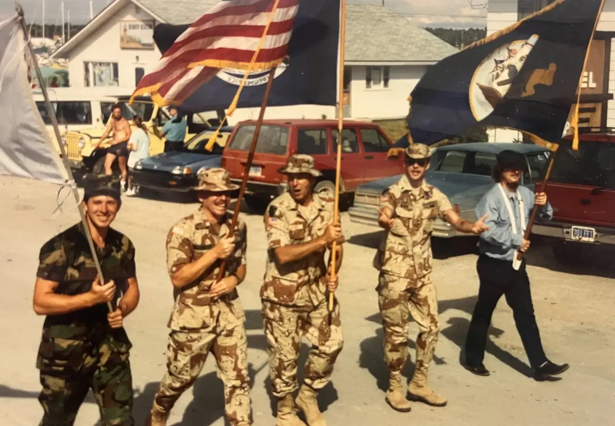 Five people in camouflage clothing and a hat march with large flags past parked cars and buildings.
