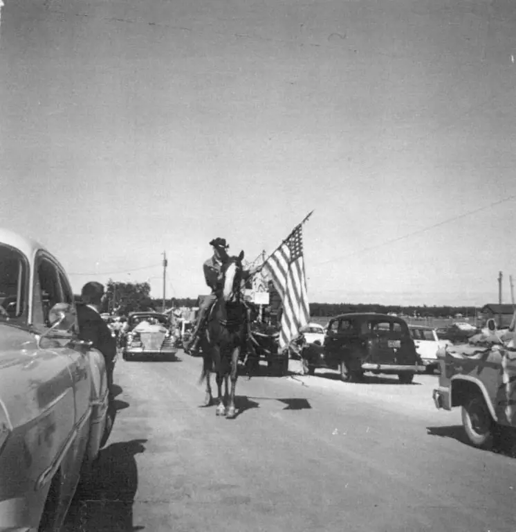 Horse-mounted rider carries an American flag down a street among parked and moving vintage cars.