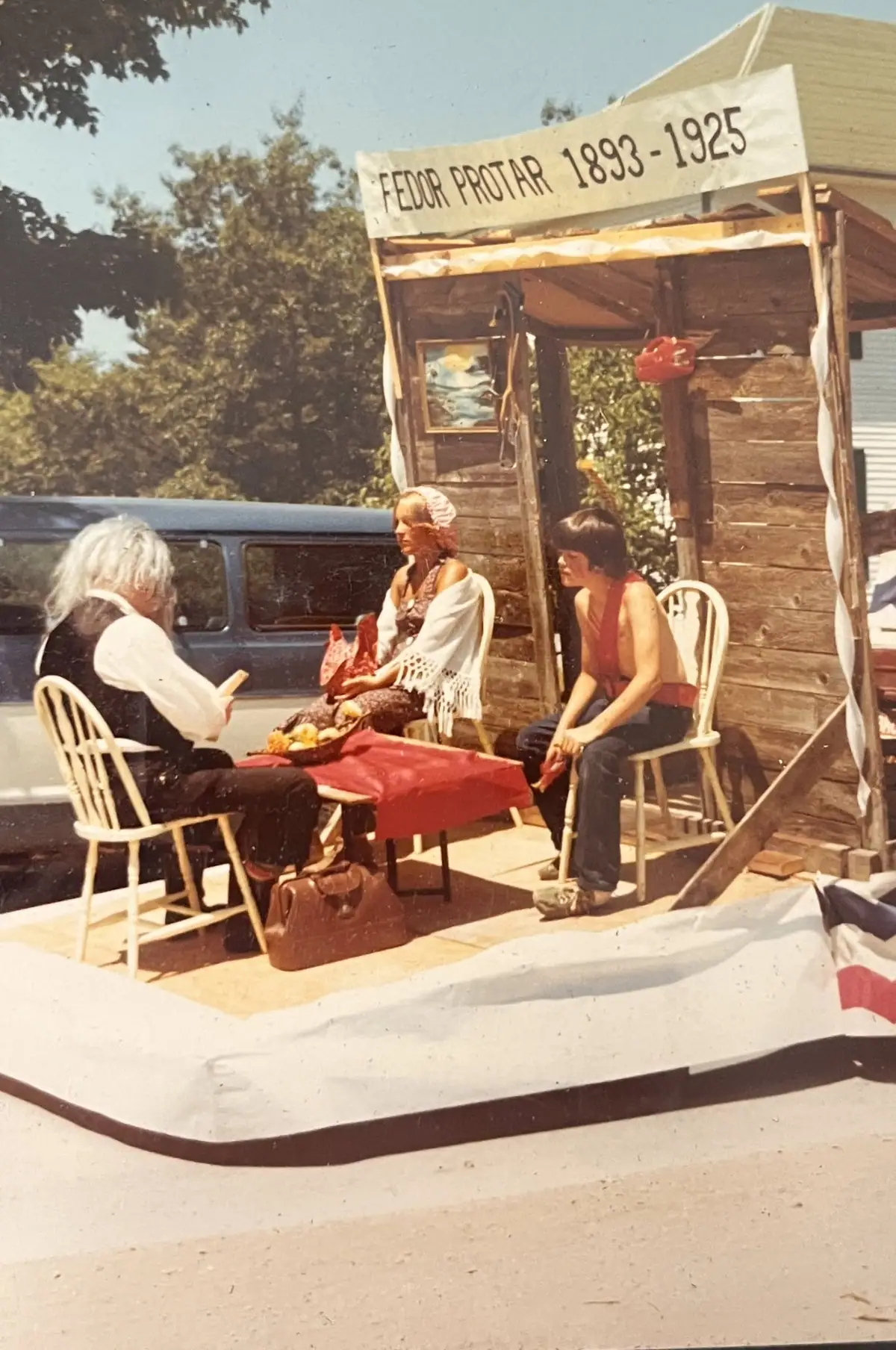 Three people sit around a table on an outdoor platform beneath a sign honoring Fedor Protar.