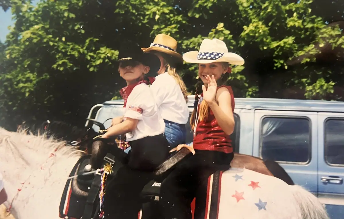 Three children in cowboy hats ride a decorated horse, with one child waving toward the camera.