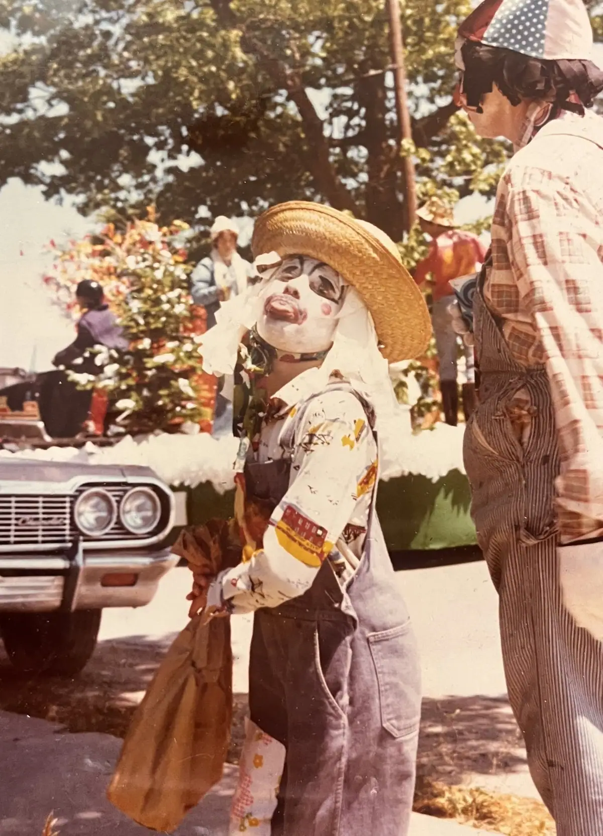 A face-painted person in a straw hat and overalls stands beside another costumed person and an old automobile.