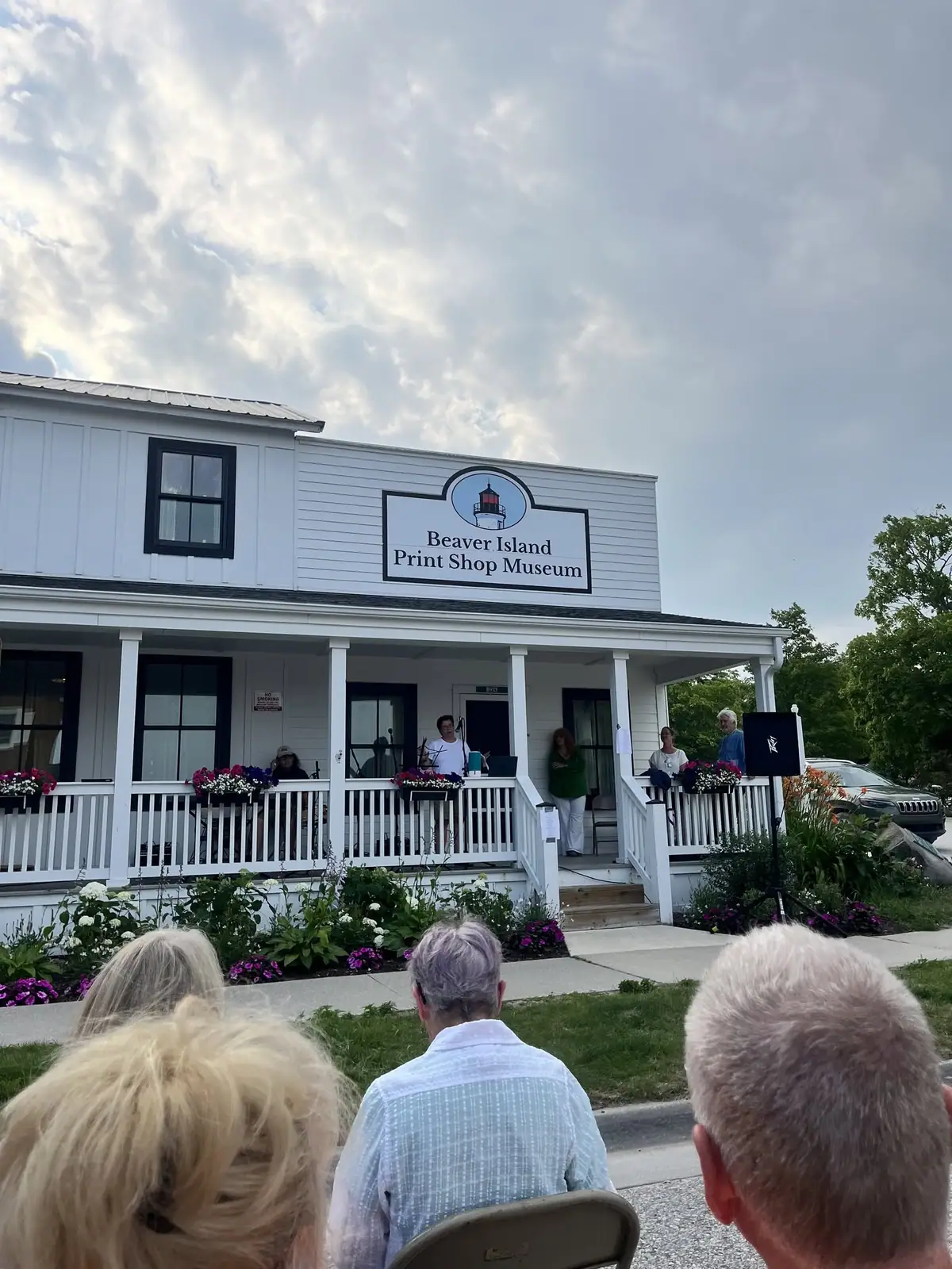 People gather outside a white building with a porch, flower boxes, and a sign reading “Beaver Island Print Shop Museum.”
