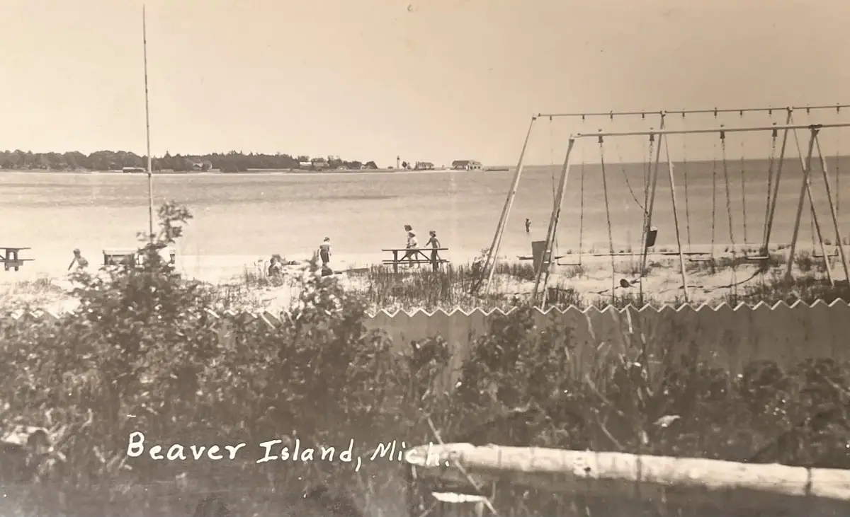 Sepia photograph of a fenced waterfront recreation area with swings, picnic tables, people, and a distant shoreline.