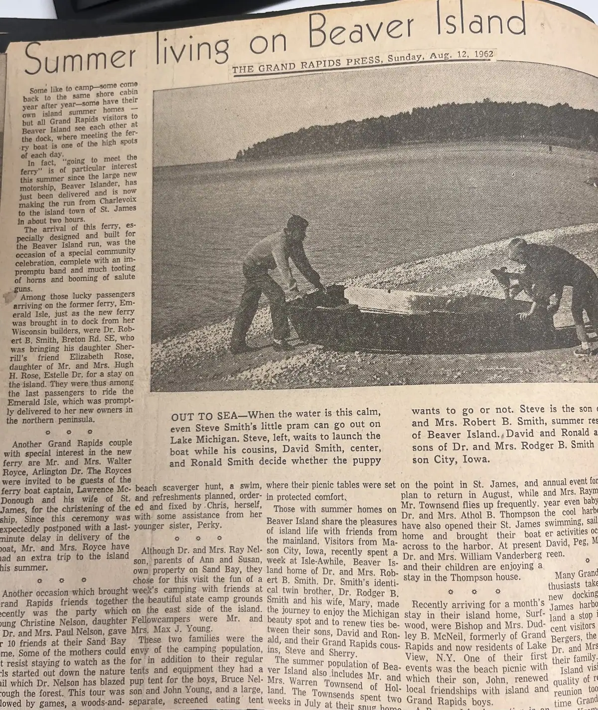 A newspaper page features a halftone photograph of three people launching a small boat from a sandy shore.