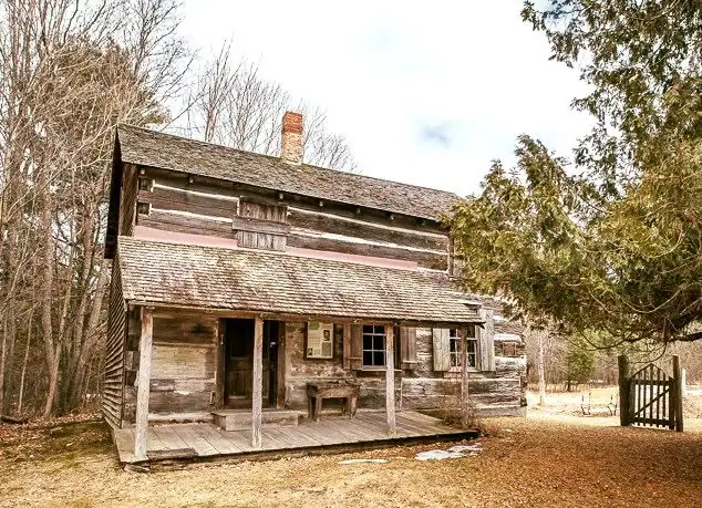 Weathered two-story log house with a covered porch, chimney, surrounding trees, and a wooden gate.