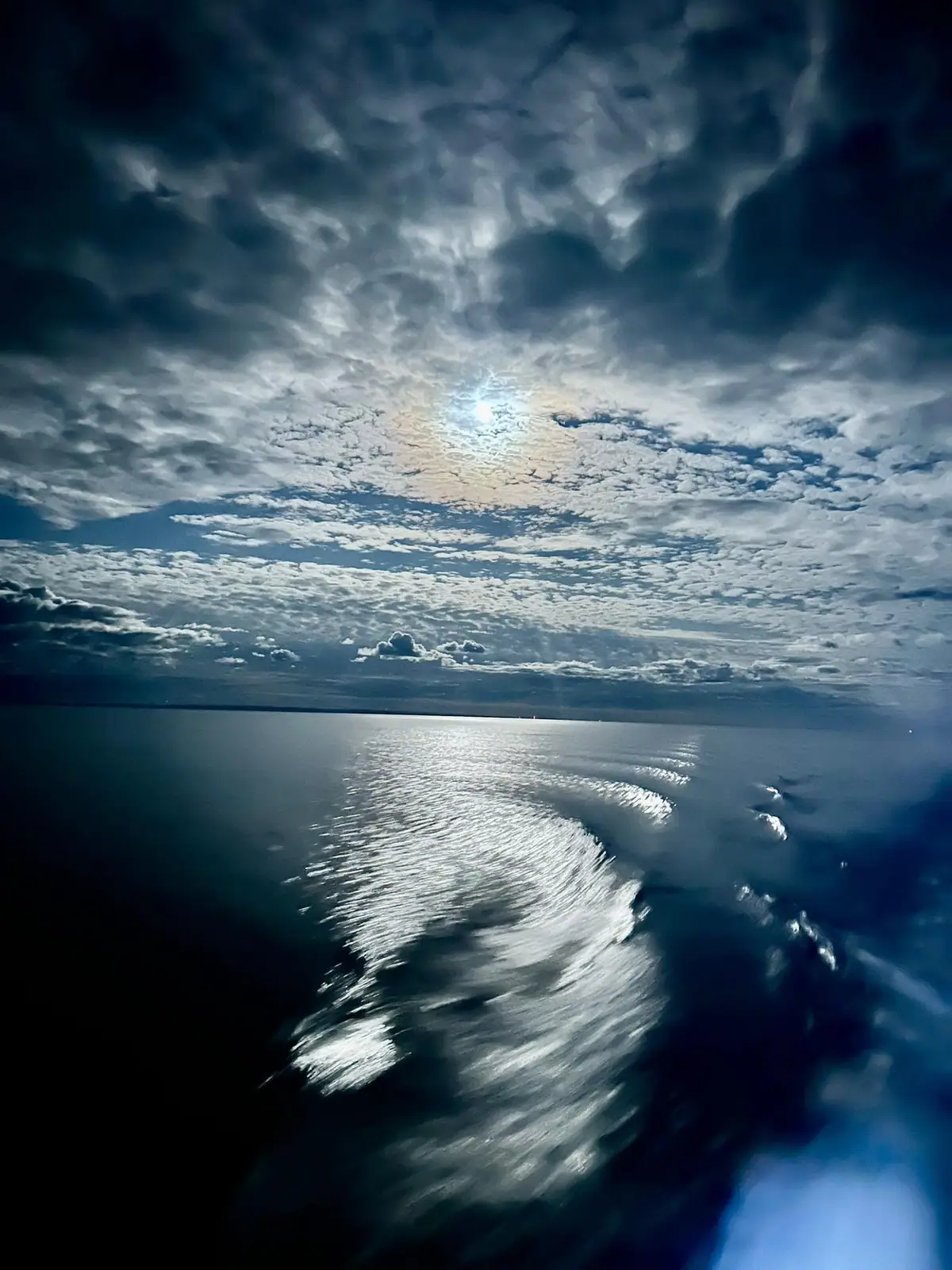 Sunlight breaks through layered clouds above open water, illuminating a broad curving wake.