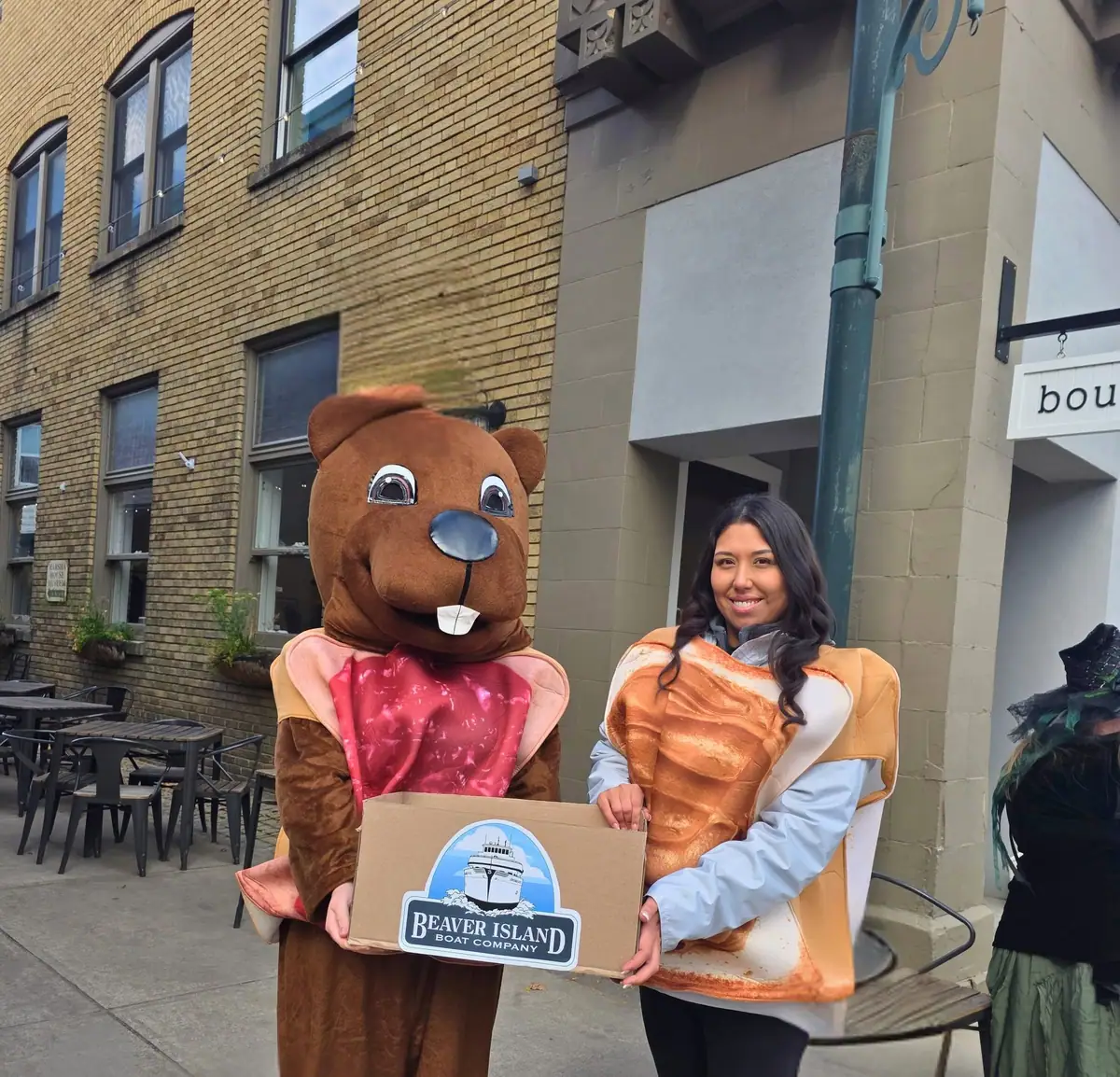 A woman and a beaver mascot hold a labeled cardboard box outside a brick commercial building.