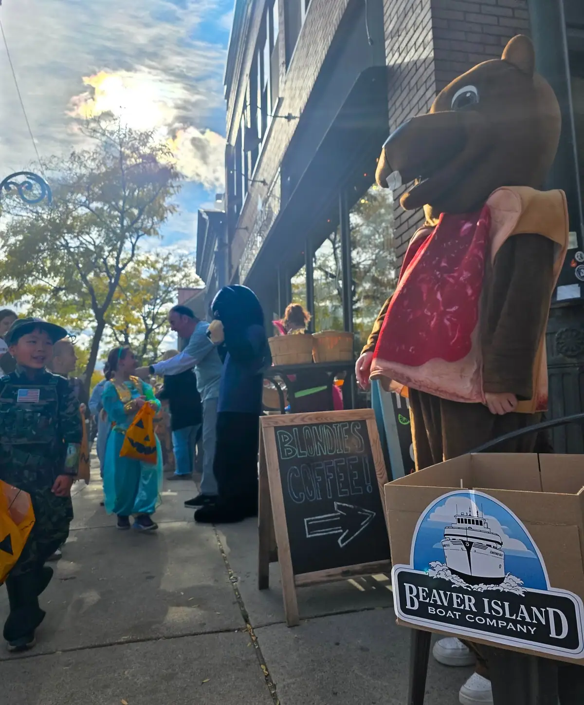 Children and adults stand beside a storefront with a large brown animal mascot, a coffee sign, and a boat-company sign.