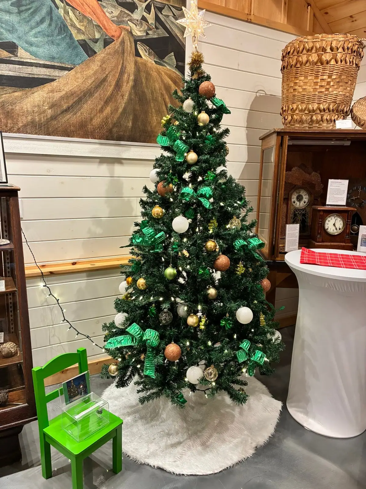 Decorated Christmas tree displayed indoors beside a green chair, clocks, a woven basket, and a large artwork.