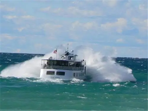 A white passenger ferry heads toward the camera through rough, foaming water beneath a partly cloudy sky.