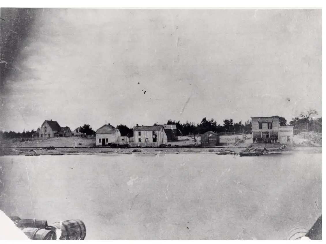 Black-and-white waterfront view of a small settlement with houses, a larger building, and barrels in the foreground.