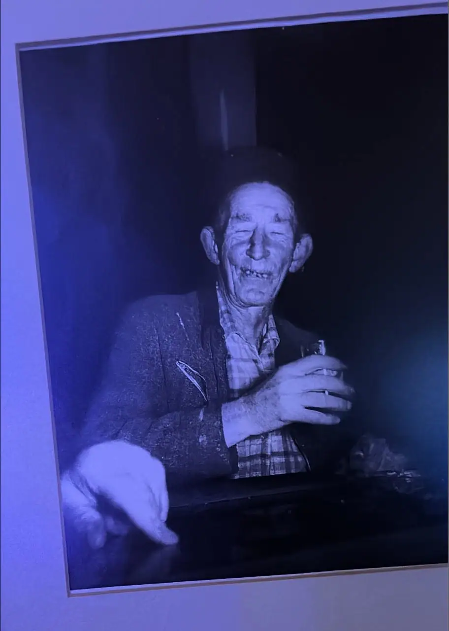 A smiling older man in a plaid shirt and jacket holds a small glass in a blue-tinted photograph.