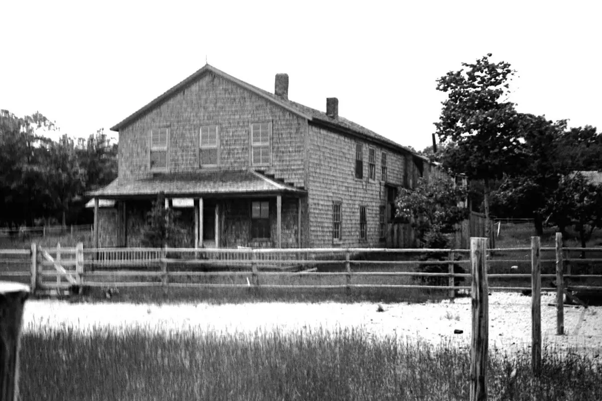 Black-and-white photograph of a two-story wooden house behind a rail fence and grassy foreground.