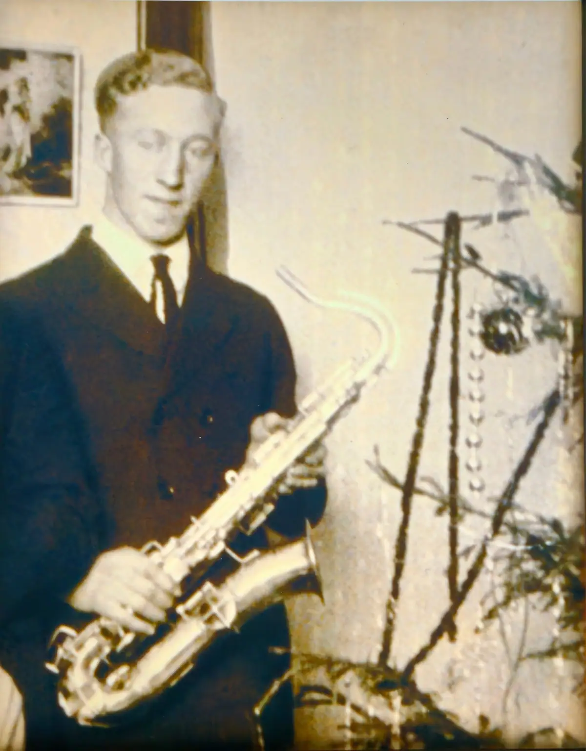 A man in a suit holds a saxophone beside a framed picture and a branch-like indoor arrangement.