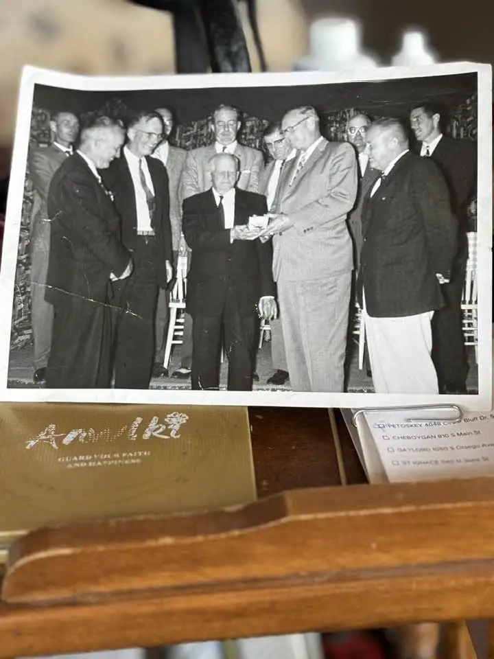 Black-and-white photograph of men in suits during a presentation, shown on a wooden surface.