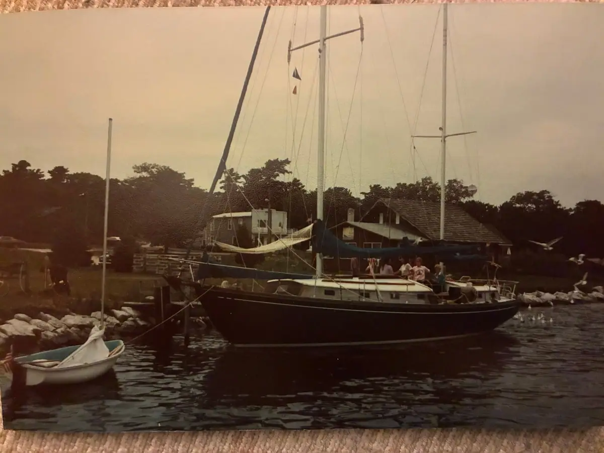 A dark-hulled sailboat with people aboard sits beside a rocky shore, with a small dinghy in the foreground.