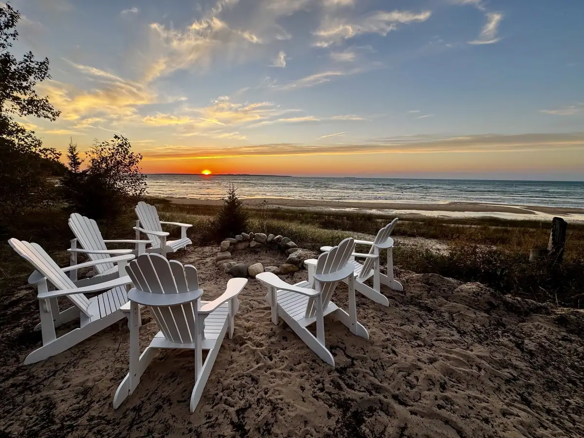 White Adirondack chairs surround a stone fire ring overlooking a beach at sunset.