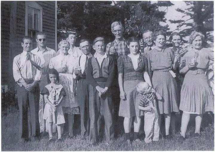 Adults and two children pose outdoors beside a wood-sided house, with several people holding drinking glasses.