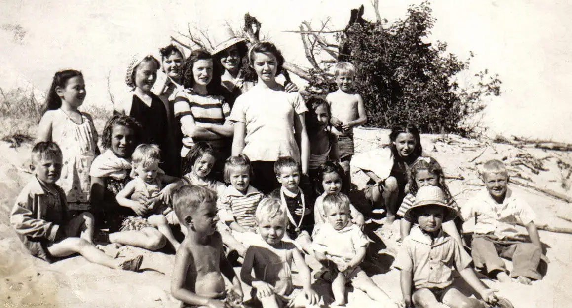 Large group of children arranged in rows on a sandy shoreline with trees and driftwood behind them.