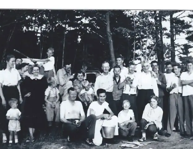 Black-and-white group portrait of adults and children gathered among trees, with a car and bucket visible.