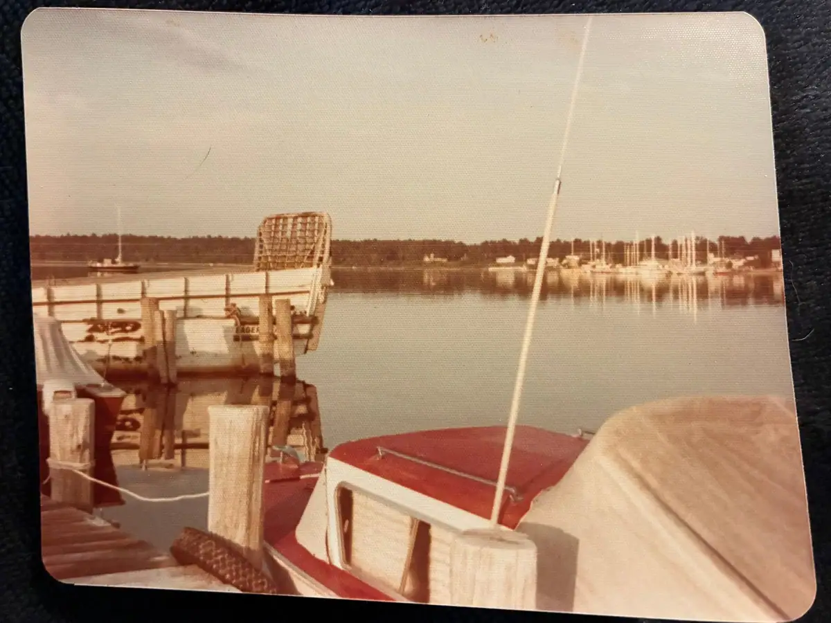 A small boat is tied to a wooden dock beside a marina, with a covered vessel in the foreground.