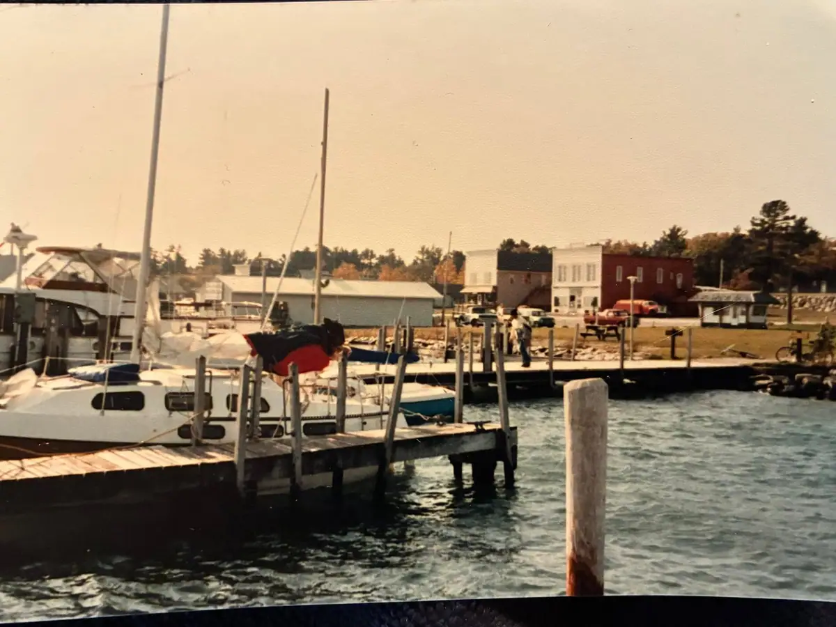 Sailboats and docks line a waterfront beside low commercial buildings and parked vehicles.
