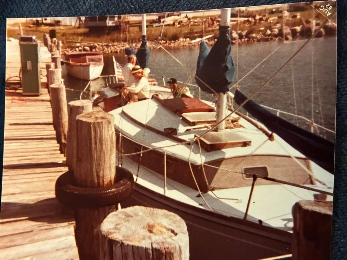 A cabin sailboat moored beside a wooden dock, with people aboard and a rocky shoreline behind.