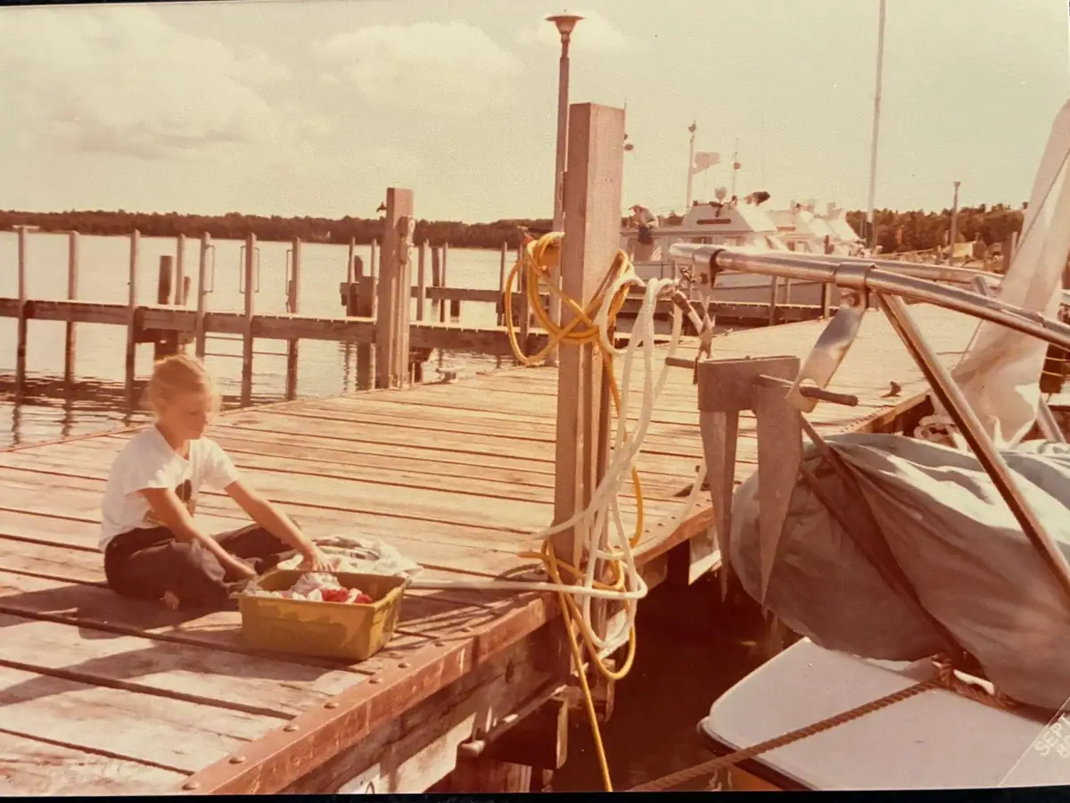 A child sorts clothing on a wooden dock beside moored boats and coiled yellow rope.