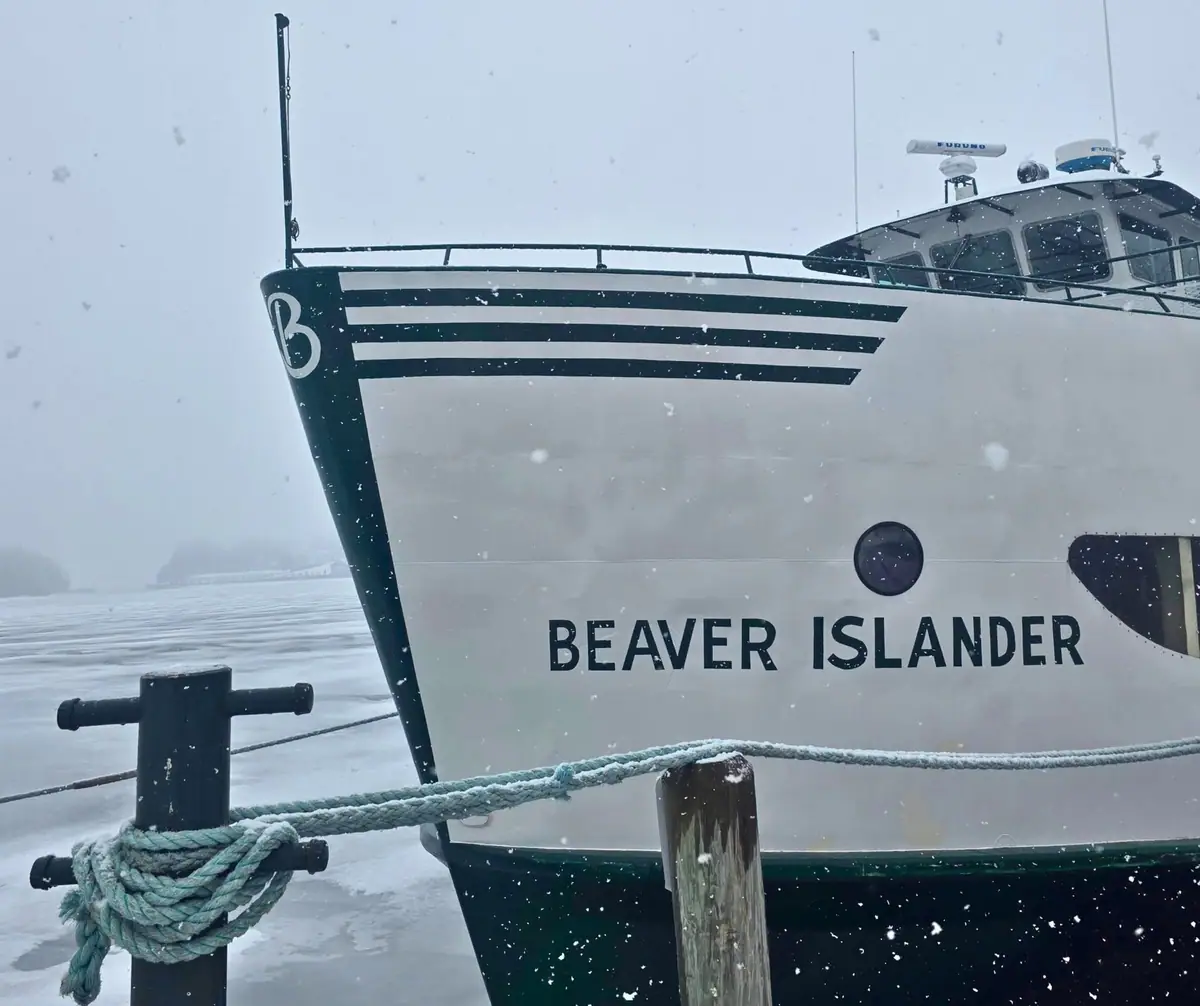 A white ferry moored at a dock during snowfall, with its hull lettering, ropes, and icy water visible.