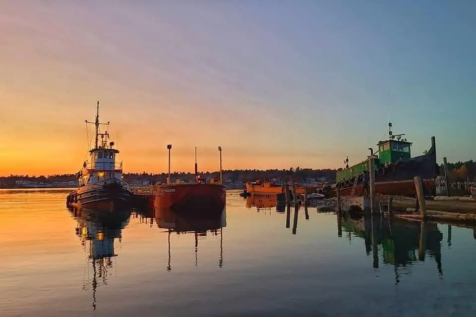 Three working vessels sit moored in calm water at sunset, reflected beside a wooden waterfront dock.