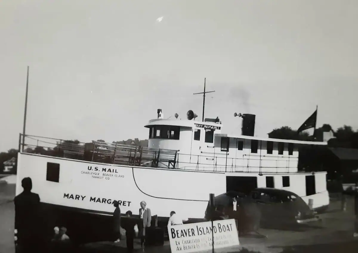 A white mail ferry named Mary Margaret beside a dock, with people, a vintage car, and a sign in the foreground.