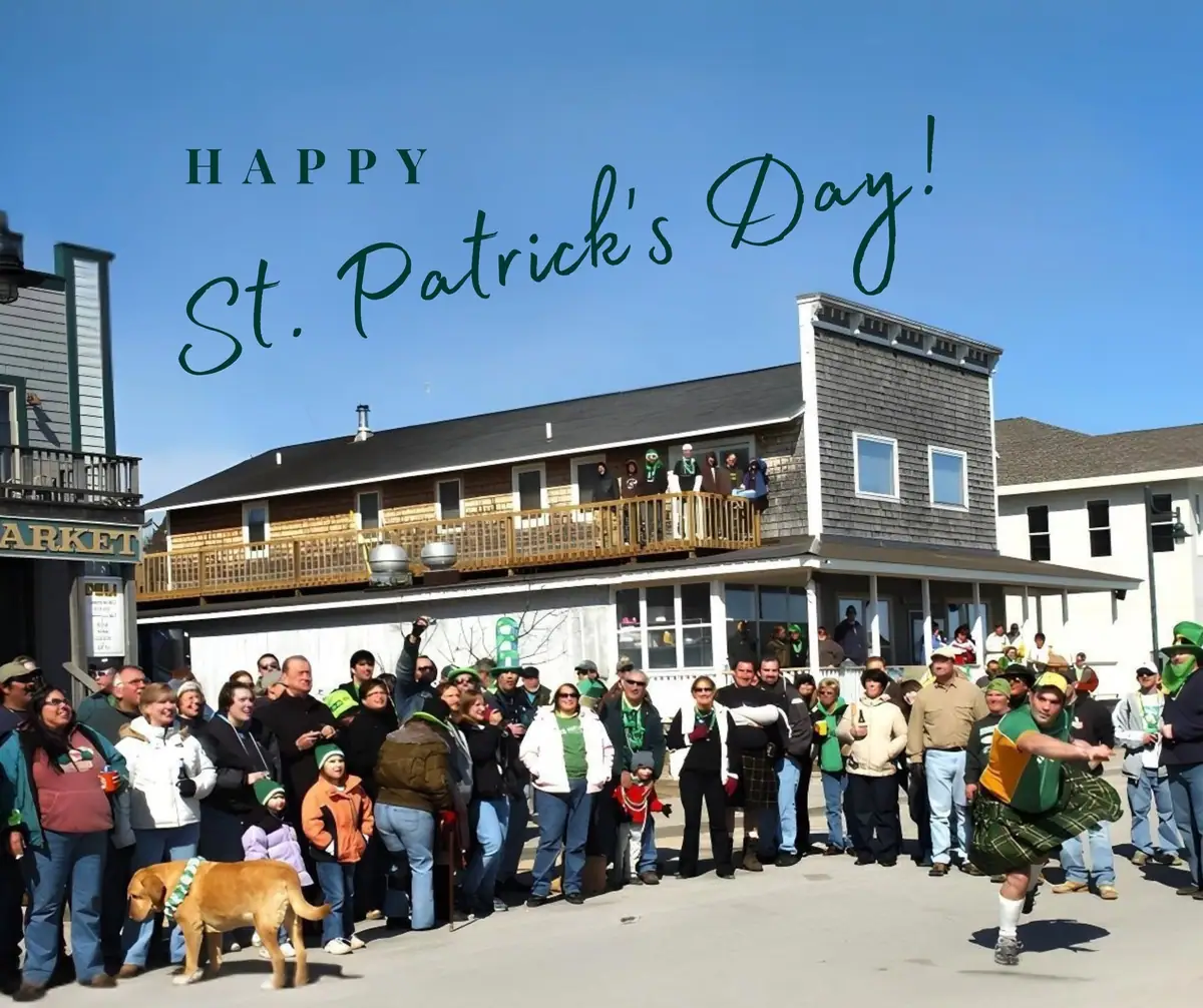 A crowd gathers outside decorated buildings for a St. Patrick’s Day celebration, with a kilted dancer and a dog in a scarf.