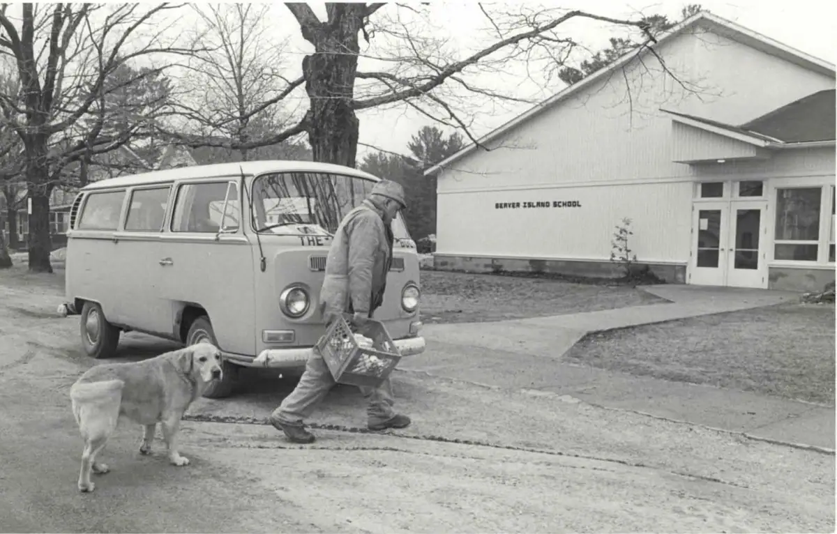 A man carries a crate beside a dog and a parked van outside a school building.