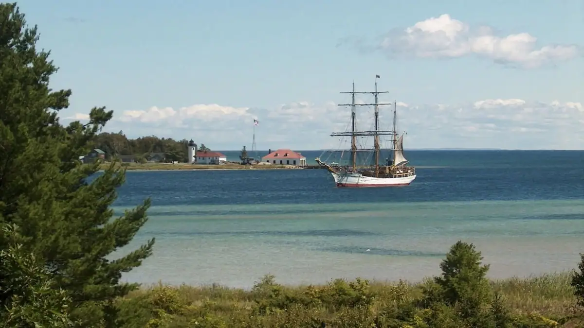 A three-masted sailing vessel passes a lighthouse and shoreline buildings beyond shallow water.