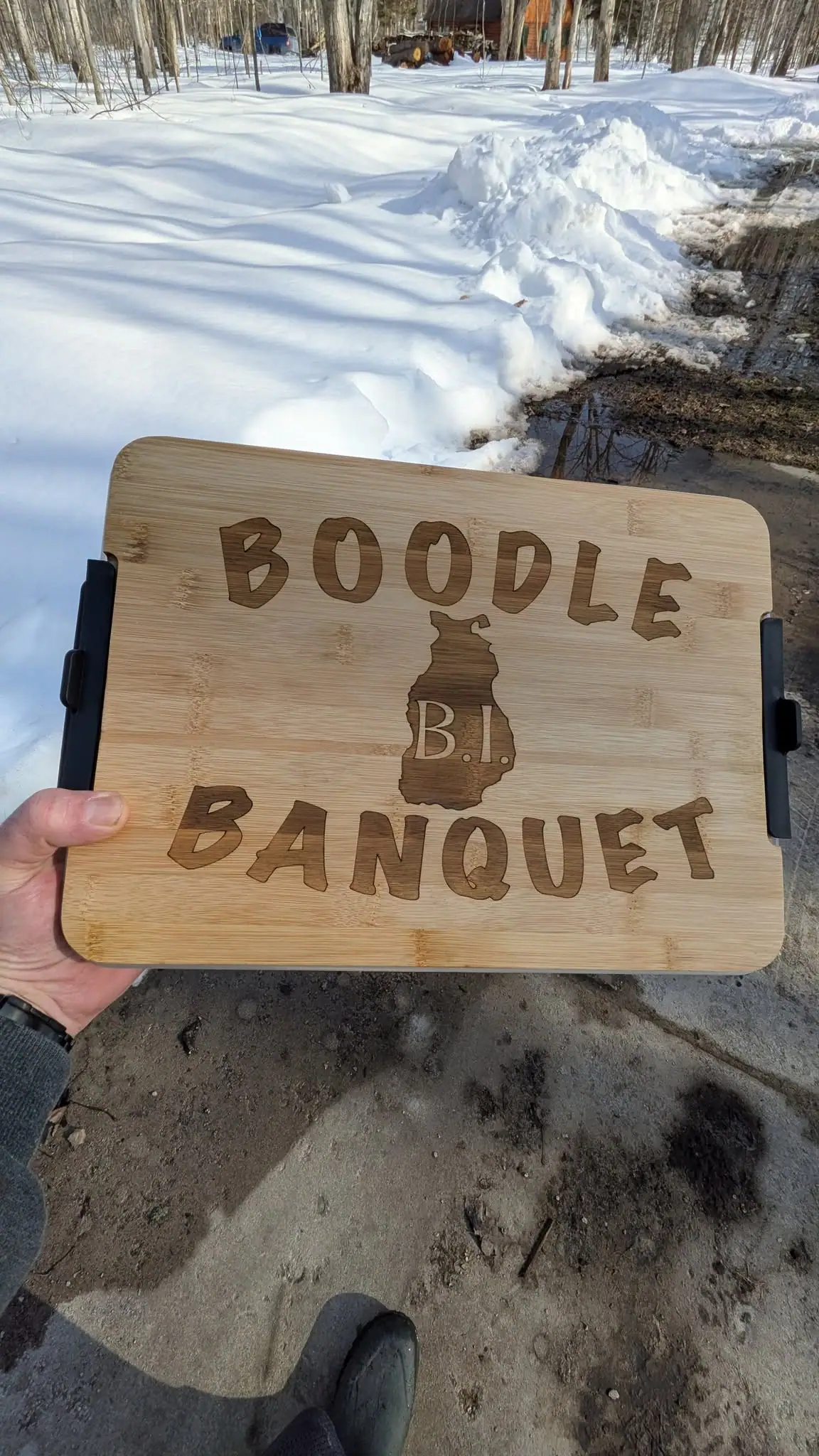 A hand holds a carved wooden board reading “BOODLE B.I. BANQUET” beside a snowy, wooded path.
