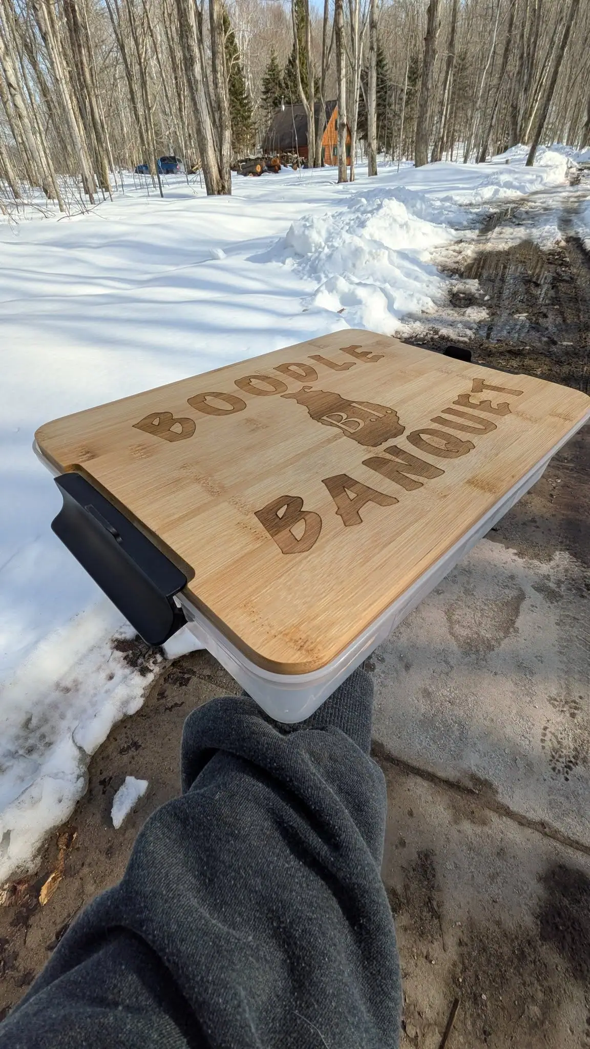A person holds a wooden-lidded container marked “BOODLE BANQUET” on a snowy wooded driveway.