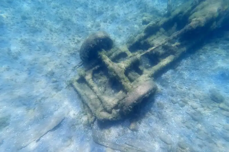 An encrusted metal vehicle frame lies on a sandy underwater bottom.