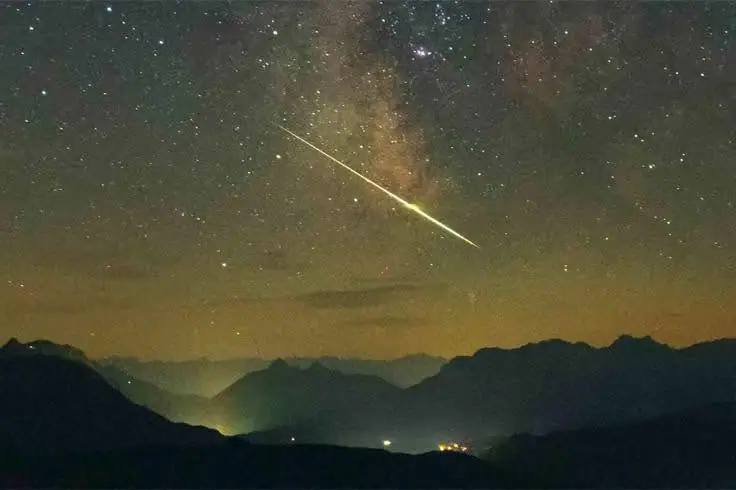 A bright streak crosses a star-filled night sky above layered mountain silhouettes and distant lights.